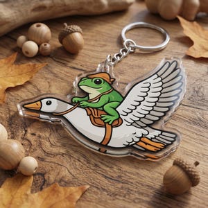 May include: A clear acrylic keychain featuring a cartoon frog riding a goose. The green frog wears an orange bandana and saddle. The goose has white wings with gray details and orange accents. A silver chain and keyring are attached.