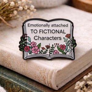 Pode incluir: Um pin de esmalte preto e branco em forma de livro aberto, com o texto "Emotionally attached TO FICTIONAL CHARACTERS". O pin é decorado com flores rosa e vermelhas e folhas verdes. O pin está em cima de dois livros.