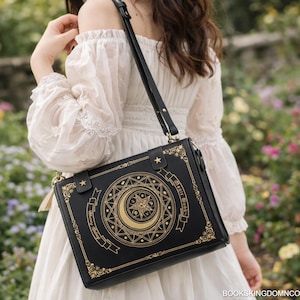Op de afbeelding: Zwarte rechthoekige crossbodytas met een gouden hemels ontwerp met een maan en ster. De tas heeft een lange zwarte riem en gouden accenten. De tekst "BOOKS KINGDOMN CO" is rechtsonder zichtbaar.