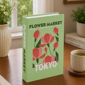 Puede incluir: Un libro verde titulado "FLOWER MARKET TOKYO" con una ilustración floral en rojo y rosa. El libro está sobre una superficie de madera, junto a una taza de té y un jarrón con flores.
