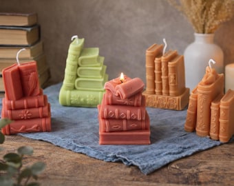 Peut inclure: Plusieurs bougies en forme de livres, de couleurs corail, vert et beige. Certaines sont allumées, révélant des détails comme des livres empilés et des pages ouvertes. Les bougies sont disposées sur un tissu bleu.
