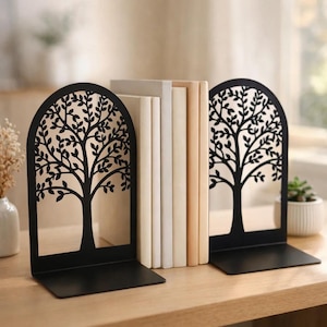 May include: Black metal tree design bookends. The bookends have a rectangular base and an arched top with a tree silhouette. A stack of books in various neutral colors sits between the bookends. The bookends are on a wooden surface.