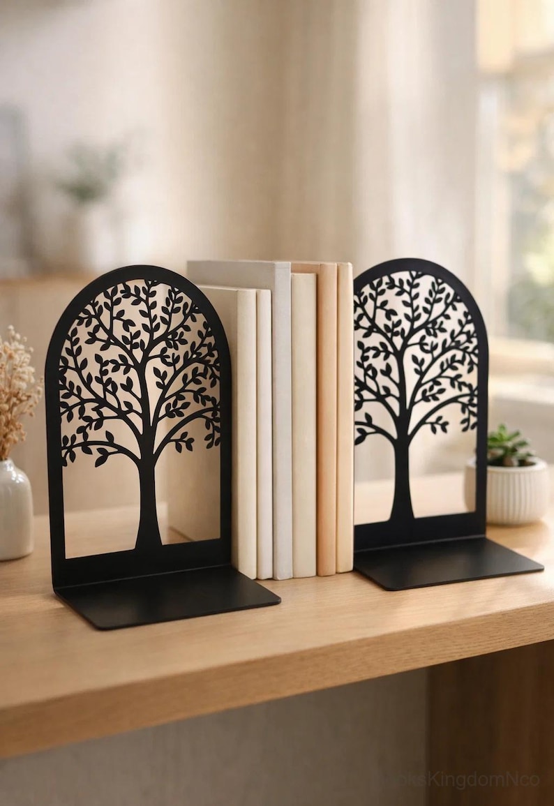 May include: Black metal tree design bookends. The bookends have a rectangular base and an arched top with a tree silhouette. A stack of books in various neutral colors sits between the bookends. The bookends are on a wooden surface.