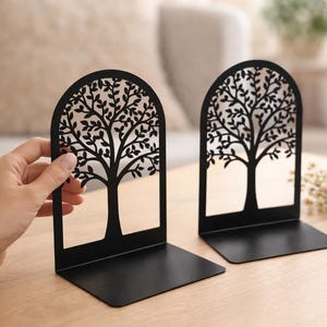 May include: Two black metal bookends with a tree design. The tree silhouette is cut out, creating an open design. The bookends are on a light-colored wooden surface. The text "BooksKingdomNco" is visible.