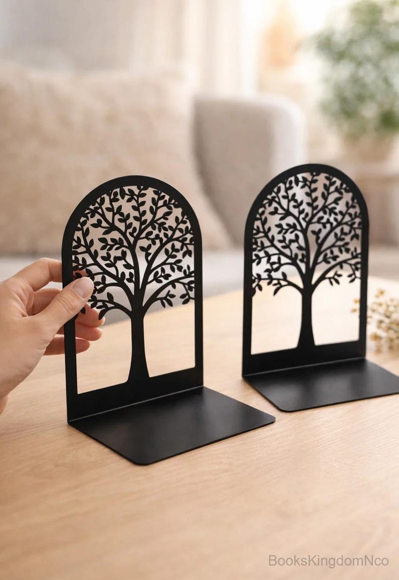 May include: Two black metal bookends with a tree design. The tree silhouette is cut out, creating an open design. The bookends are on a light-colored wooden surface. The text "BooksKingdomNco" is visible.