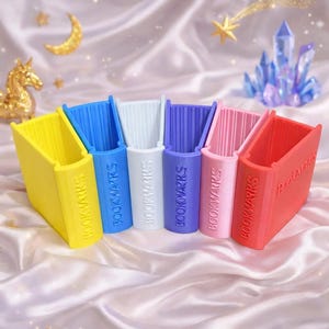 Peut inclure: Ensemble de marque-pages colorés en forme de livre, jaunes, bleus, blancs, violets, roses et rouges. Chaque marque-page porte l'inscription "BOOKMARKS" sur un fond satiné avec des décorations célestes.