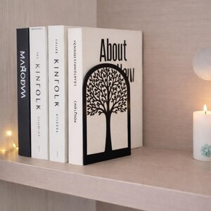May include: A black metal tree-shaped bookend holds several books on a shelf. The books have white and black covers with titles like "About" and "Kinfolk". A lit candle and decorative lights are in the background.