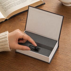 Könnte beinhalten: Ein buchförmiger Safe mit Zahlenschloss. Der Safe ist geöffnet und zeigt einen grauen Innenraum. Das Buch hat weiße Seiten und einen schwarzen Einband. Eine Hand dreht das Kombinationsrad am Schloss.
