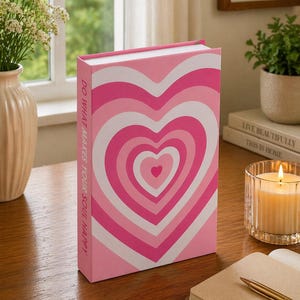 May include: A pink journal featuring a heart design in varying shades of pink and white. The spine displays the text "DO WHAT MAKES YOUR SOUL HAPPY". The journal rests on a wooden surface, alongside a lit candle and a notebook.