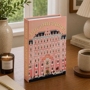 以下が含まれることがあります： 背表紙と表紙に「GRAND BUDAPEST HOTEL」の文字が入ったピンク色の本。表紙には、複数の窓と黒い屋根があるピンク色の建物の詳細なイラストが描かれています。白い車と人々が下部に描かれています。