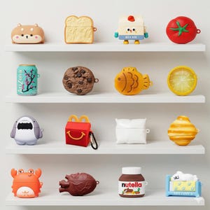 Op de afbeelding: Een verzameling novelty AirPod-hoesjes in verschillende vormen, waaronder een hond, toast, melkpak, tomaat, Arizona-blikje, koekje, vis, citroenschijfje, haai, McDonald's-doos, kussen en een krab. De hoesjes worden op witte planken getoond.