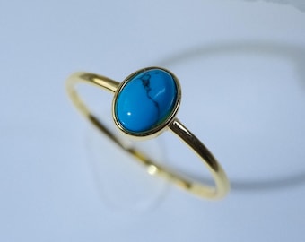 May include: A gold ring featuring a small, oval-shaped turquoise gemstone set in a smooth bezel. The band is thin and polished, showcasing a bright blue stone with subtle dark veining, positioned near a white flower with a yellow center.