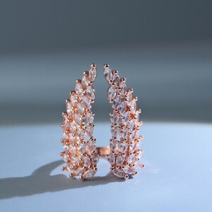 May include: A rose gold ring featuring two symmetrical wing-shaped sections adorned with numerous marquise-cut clear stones. The design consists of two separate, curved rows of sparkling crystals that meet at the base, creating an open wing motif.