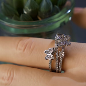 May include: A silver-toned ring featuring two butterfly motifs and a single round clear stone. The band is split into two thin, parallel sections, both encrusted with small, sparkling clear stones that catch the light, creating a delicate appearance.
