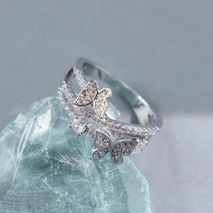 Puede incluir: Un anillo de tono plateado con dos motivos en forma de mariposa incrustados con pequeñas piedras claras. La banda está dividida en dos filas, ambas pavimentadas con diminutas piedras claras, e incluye una piedra redonda clara entre las mariposas.