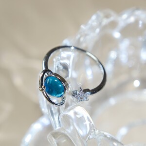 May include: A silver-toned open ring featuring a round, translucent blue planet-shaped charm with a thin encircling ring on one end and a small, five-pointed star encrusted with tiny clear stones on the other end of the adjustable band.