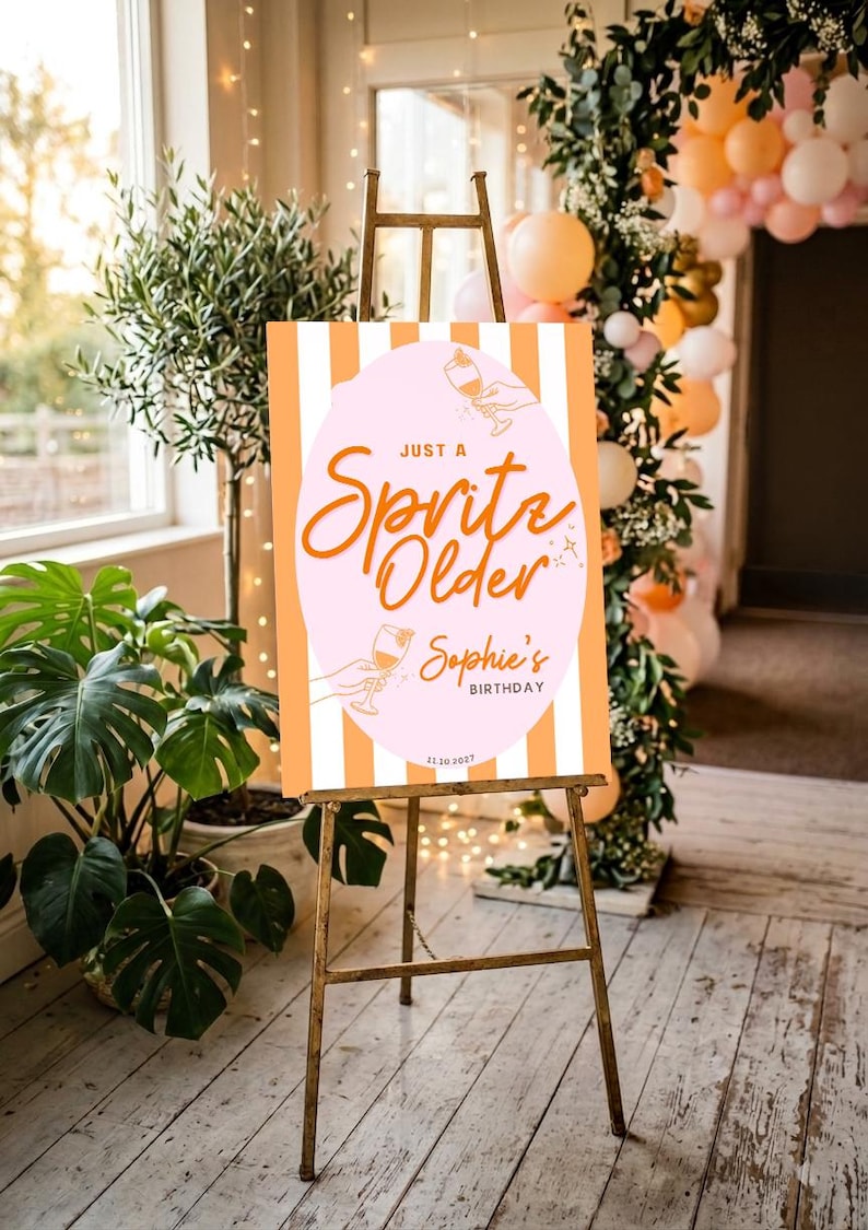 May include: A party sign on a wooden easel with the text "Just a Spritz Older Sophie's Birthday" in orange script. The sign has a pink oval with a cocktail illustration and orange and white vertical stripes.