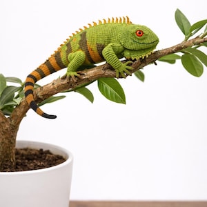 May include: A handcrafted, green crochet iguana with orange and brown stripes, perched on a tree branch with green leaves. The iguana has red eyes and is set against a white background. The tree is in a white pot.