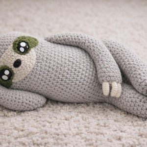May include: A handmade crochet sloth toy in light gray, lying on a cream-colored surface. The sloth has a beige face with large black and white eyes, green eye patches, and cream-colored claws.