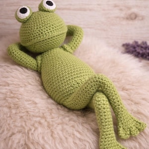 Pode incluir: Um brinquedo de sapo em crochê na cor verde-clara com cabeça arredondada, olhos grandes brancos e pretos e corpo rechonchudo. O sapo está em posição reclinada com os braços atrás da cabeça e as pernas cruzadas, apresentando pontos de crochê texturizados.