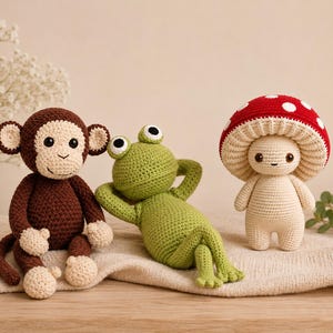 Puede incluir: Tres juguetes de ganchillo: un mono marrón con manos y pies color crema, una rana verde tumbada con las manos detrás de la cabeza y una figura de seta color crema con sombrero rojo de puntos blancos y ojos oscuros.