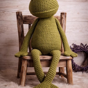 Könnte beinhalten: Eine handgefertigte, olivgrüne Häkelfroschpuppe mit großen, runden Augen. Der Frosch sitzt auf einem kleinen Holzstuhl, seine langen Beine sind ausgestreckt. Der Körper des Frosches ist strukturiert, und die Augen haben weiße und schwarze Details.