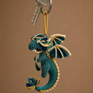 Puede incluir: Un llavero de dragón tejido a ganchillo en colores azul verdoso, azul oscuro y crema. El dragón tiene ojos grandes y oscuros, alas detalladas y una cola enroscada. Está unido a una anilla de metal plateado con la palabra KALE grabada.