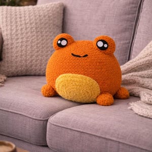 May include: An orange frog-shaped pillow with a yellow belly and black eyes sits on a gray couch. The plush pillow has a smiling face and small, round feet. The background includes a textured pillow and a soft blanket.
