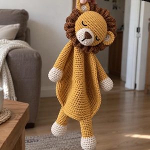 Può includere: Una copertina di sicurezza a forma di leone realizzata all'uncinetto, con corpo giallo oro, criniera marrone scuro e zampe color crema. La testa del leone presenta occhi chiusi ricamati e un naso marrone. La coperta è circolare e testurizzata.