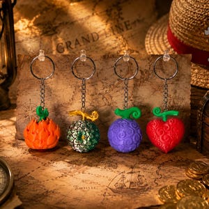 Könnte beinhalten: Vier farbenfrohe, fruchtförmige Schlüsselanhänger mit silbernen Ketten und Ringen. Die Früchte sind orange, grün, lila und rot, jede mit einem einzigartigen Design. Die Schlüsselanhänger werden auf einer Vintage-Karte mit Kompass und Schatzkiste im Hintergrund präsentiert.