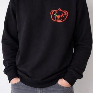 Könnte beinhalten: Schwarzes Sweatshirt mit Rundhalsausschnitt und einem rot-grünen, gestickten Bärenmotiv auf der Brust. Der Bär hat ein Dollarzeichen als Auge und ein Kreuz als Mund. Lässiger Stil.