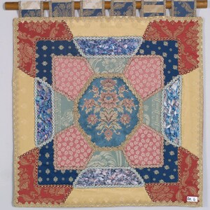 Può includere: Un arazzo decorativo con un design patchwork. Il pezzo quadrato presenta un medaglione floreale blu centrale circondato da tessuti a motivi rosa, blu e rossi. Rifiniture dorate e un'asta di legno con passanti in tessuto completano il pezzo.