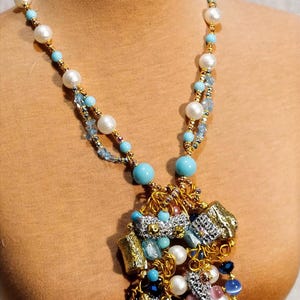 Peut inclure: Un collier fantaisie avec une chaîne dorée, de grosses perles blanches, et des perles turquoise. Le pendentif est composé de pièces métalliques argentées et dorées, de pierres bleues et roses, et d'autres fausses perles.