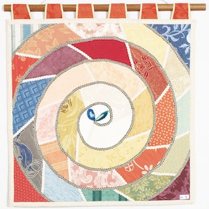 Può includere: Un arazzo da parete colorato a forma di spirale. Il design presenta toppe di tessuto in tonalità di rosso, giallo, blu e verde, bordate da un cordoncino bianco e nero. È montato su un'asta di legno con anelli di nastro arancione.