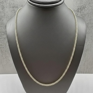 May include: A delicate, silver-toned tennis necklace displayed on a gray mannequin. The necklace features a continuous line of small, square-cut, clear gemstones. The stones are closely set, creating a sparkling, elegant appearance.