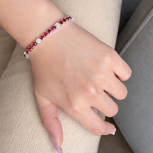 May include: A tennis bracelet with alternating red and clear gemstones. The bracelet, set in silver-coloured metal, is displayed on a person's wrist. The gemstones are likely rubies and diamonds.