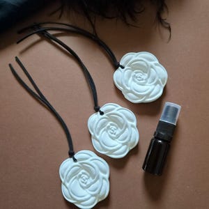Puede incluir: Tres adornos de cerámica blanca con forma de rosa, cada uno con un cordón negro para colgar, acompañados de un pequeño frasco pulverizador de vidrio ámbar oscuro con una tapa de plástico transparente.