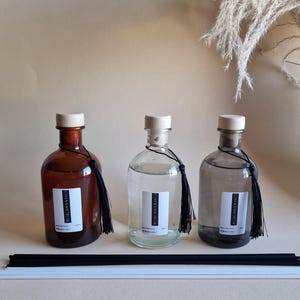 Op de afbeelding: Drie glazen diffuserflessen met houten doppen en zwarte kwastjes. De flessen zijn gevuld met vloeistof en hebben witte etiketten. Zwarte en witte diffuserstokjes liggen voor de flessen. De flessen zijn in verschillende tinten bruin, helder en grijs.