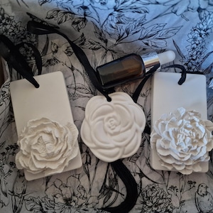 Könnte beinhalten: Drei weiße Keramikanhänger mit schwarzen Bändern und geprägten Blumendesigns, zusammen mit einer kleinen braunen Glasflasche mit einer Pipette. Die Anhänger zeigen ein Rosen- und Pfingstrosen-Design. Der Hintergrund ist ein Stoff mit floralem Muster.