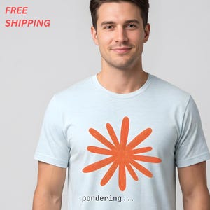 May include: A light blue short-sleeved t-shirt featuring a large, orange, multi-petaled flower graphic centered on the chest. Below the graphic, the word "pondering..." is printed in a small, black, sans-serif font on the front of the shirt.