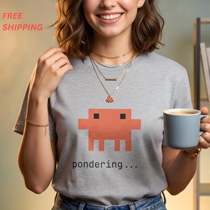 May include: A light gray short-sleeve t-shirt featuring a pixelated orange alien graphic and the text "pondering..." printed in black. The shirt is worn with layered gold necklaces and a light blue ceramic mug is held in one hand.