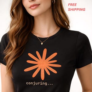 May include: Black t-shirt featuring an orange burst design with the text "conjuring...". The shirt has short sleeves and a crew neck. A gold necklace with a heart pendant is visible.