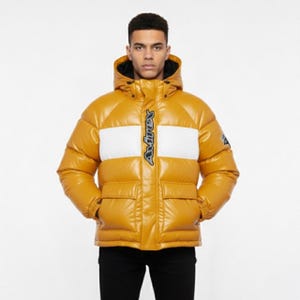 May include: Mustard yellow puffer jacket with a white stripe across the chest. The jacket has a hood, two front pockets, and the word "Avirex" in black script down the center. The jacket is worn by a person wearing black pants.