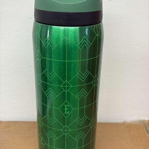 May include: A green insulated tumbler with a black lid and a geometric pattern. The tumbler has a metallic finish and a flip-top lid. The design includes the letter "E" within the pattern. The tumbler is designed to keep beverages hot or cold.