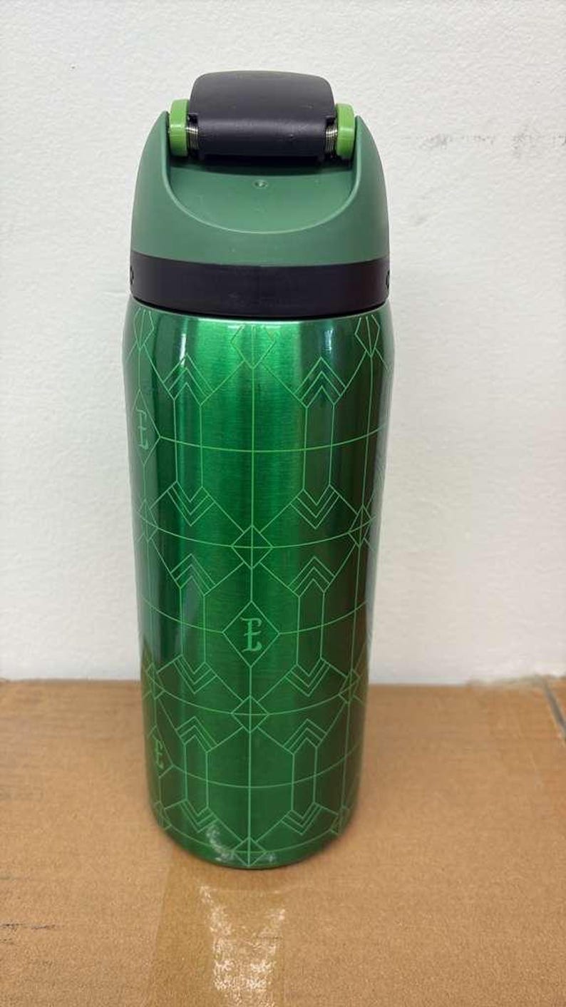 May include: A green insulated tumbler with a black lid and a geometric pattern. The tumbler has a metallic finish and a flip-top lid. The design includes the letter "E" within the pattern. The tumbler is designed to keep beverages hot or cold.