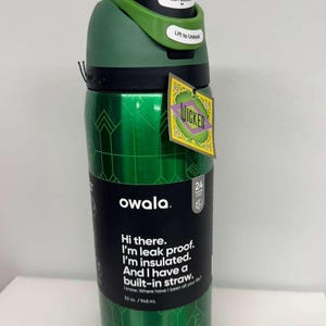 May include: Green Owala water bottle with a black lid and a green handle. The bottle has a geometric pattern and the word "WICKED" on a tag. Text on the bottle reads "Hi there. I'm leak proof. I'm insulated. And I have a built-in straw."