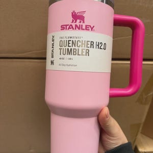 May include: A pink Stanley Quencher H2.0 tumbler with a matching handle and lid. The tumbler has the Stanley logo and product information printed on the front. It holds 1.18 L.