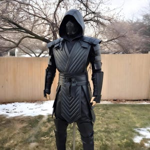 Puede incluir: Un disfraz de cosplay negro con una parte superior con capucha y máscara, armadura para los hombros y una túnica acolchada. El disfraz incluye protectores de brazos, un cinturón y pantalones a juego. El atuendo se exhibe en un maniquí al aire libre.