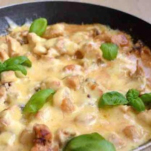 May include: A skillet filled with a creamy, cheesy chicken dish, garnished with fresh green basil leaves. The chicken pieces are golden brown, and the sauce is a rich, yellow color. A home-cooked meal.