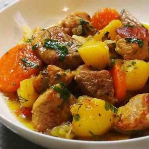 May include: A close-up shot of a white bowl filled with a hearty stew. The stew features chunks of cooked meat, bright orange carrots, and yellow potatoes, all glistening in a savory sauce. Fresh green herbs are sprinkled throughout.